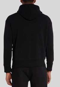 Uomo che indossa una felpa nera semplice e pantaloni neri, visto da dietro su uno sfondo grigio uniforme.