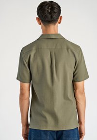 Koszula z krótkim rękawem w oliwkowym odcieniu, wykonana z teksturowanej tkaniny. Charakteryzuje się płaskim kołnierzykiem i zaszewką z tyłu, co zapewnia swobodę ruchu.