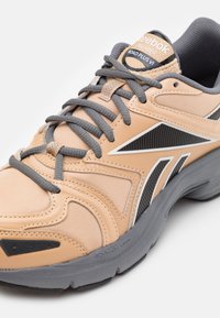 Baskets Reebok Premier Road Plus VI beige et grises avec semelle texturée, lacets gris foncé et logo latéral noir et blanc.