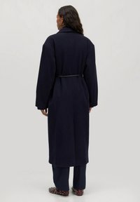 Marineblauw wollen jas met lange mouwen, ceintuur in de taille, brede kraag en zijzakken, gecombineerd met donkere broeken en gestifte schoenen.