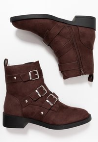 Botas de tobillo de ante marrón con tres correas metálicas de hebilla, cremallera lateral y un tacón bajo negro. Textura suave, diseño de punta afilada.