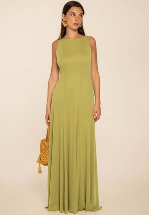 Mujer con un vestido maxi sin mangas verde oliva sosteniendo un bolso tejido amarillo, de pie frente a un fondo neutro.