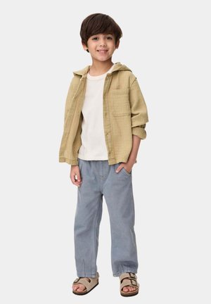 Beige Texturhemd mit Kapuze, weißem T-Shirt, hellblauen Jeans und braunen Sandalen. Das Hemd hat zwei Brusttaschen und hochgekrempelte Ärmel.