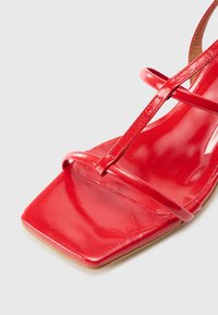 Sandale en cuir rouge avec un bout carré, dotée de fines lanières croisées et d'une finition lisse et brillante. La semelle est de couleur marron clair et plate.