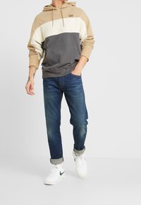 Sudadera con capucha en tonos marrón claro, crema y gris oscuro, con bolsillo canguro y logo bordado. Combinada con jeans azules y zapatillas blancas.