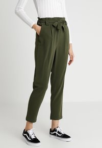 Pantalones verde oliva de cintura alta con detalle de lazo, que presentan bolsillos laterales y piernas ajustadas, combinados con zapatillas negras y blancas.