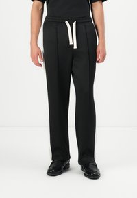 Black trousers with a relaxed fit, featuring an elastic waistband and white drawstring. Side stripe detail adds contrast. Smooth texture.