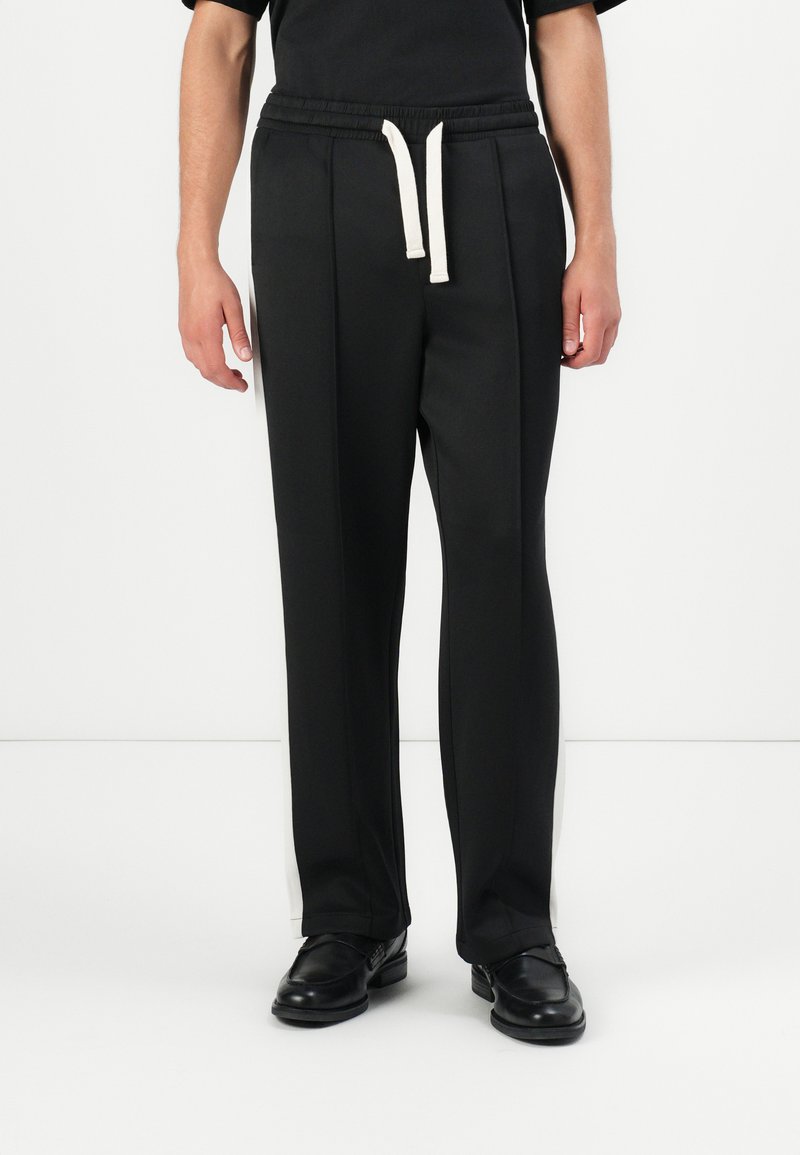 Black trousers with a relaxed fit, featuring an elastic waistband and white drawstring. Side stripe detail adds contrast. Smooth texture.