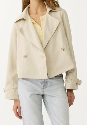 Femme portant un trench-coat beige court par-dessus un haut jaune boutonné et un jean bleu clair, debout avec une main détendue sur le côté.