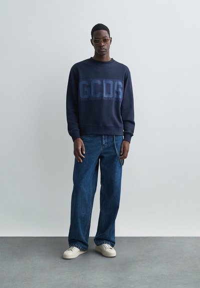Marinblå sweatshirt med "GCDS"-logotyp i en mörkare nyans, matchad med vida blå denimjeans och beige sneakers. Minimalistisk design och textur.