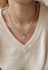 Zilveren halsketting met drie strengen: een gladde ketting, een gestructureerde ketting en een hanger met een hartvormige bedel, gedragen over een beige trui.