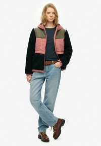 Fleece gevoerde jas met een roze en olijfkleurig blokontwerp, ritssluiting aan de voorkant en een zwarte, textuurlijke body. Gecombineerd met blauwe jeans en bruine laarzen.