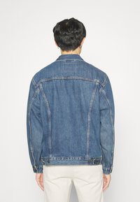 Chaqueta de mezclilla en azul medio, con un corte clásico, costuras cosidas, cierres de botón en los puños y detalles en el yugo trasero.