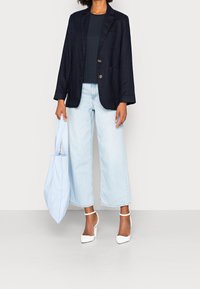 Kvinna i marinblå kavaj, svart topp, ljust blå vida jeans, vita klackar, som håller en blå och vit randig tote-väska.