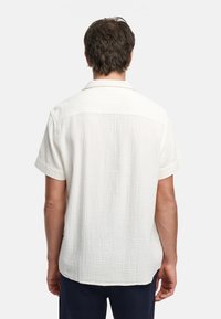 Camisa de manga corta de color beige claro, fabricada en algodón texturizado, con un cuello plano y un corte holgado. La vista trasera muestra un sutil patrón de cuadrícula.