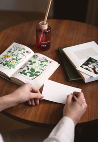 Une table en bois supporte un livre ouvert avec des illustrations botaniques, un vase en verre rouge contenant des roseaux, et une carte blanche en cours d'écriture manuscrite.