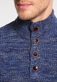Strickpullover mit hohem Kragen in einem blau-grauen Melange-Muster, verziert mit vier dunklen Knöpfen mit kontrastierenden roten Rändern.
