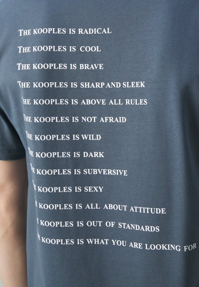Tamsiai mėlynas medvilnės marškinėliai su balta tekstine fraze, apibūdinančia "The Kooples", išdėstyta vertikaliai su skirtingais eilučių aukščiais.