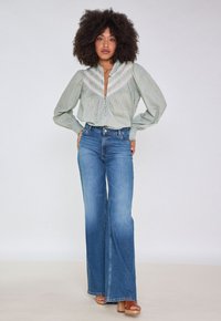 Blusa verde chiaro con dettagli ricamati, maniche ampie e scollatura profonda a V, abbinata a jeans blu a gamba larga a vita alta e tacchi marroni.