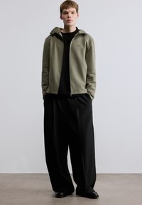 Ζακέτα με φερμουάρ σε ελιά πράσινο χρώμα και κουκούλα, μαύρο μπλουζάκι με λαιμόκοψη crew από κάτω, και φαρδιά μαύρη παντελόνα· ομαλό ύφασμα, με ελάχιστο λογότυπο στη ζακέτα.