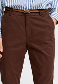 Braune Baumwollhosen mit geradem Schnitt, vorderen Taschennähten und einem braunen Gürtel mit einer silbernen Schnalle. Sichtbares Hemd mit horizontalen Streifen.