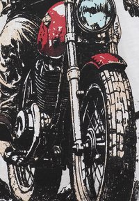 Ruota anteriore di una motocicletta con battistrada spesso e parafango rosso, gambe del pilota in pantaloni beige e fanale tondo visibile in primo piano.