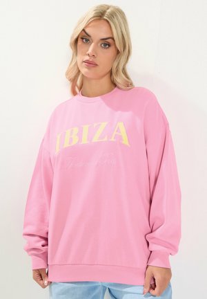 Blonde Frau trägt einen locker sitzenden pinkfarbenen Pullover mit dem Schriftzug "IBIZA Balearisches Meer" und steht vor einem weißen Hintergrund.