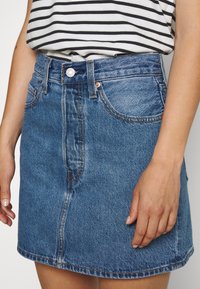 Kvinna som bär en blå denimkjol och en vit och svart randig ärmlös topp, visar framsidan av midjan och händerna.