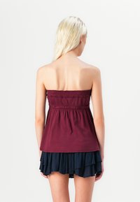 Bordeaux strapless top met smocktextuur, gecombineerd met een gelaagde zwarte roklengte met ruffles. Beide kledingstukken zijn gemaakt van zachte, lichte stof.