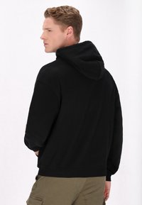 Sort hætte sweatshirt lavet af blødt stof, med lange ærmer, ribbede manchetter og en kængurulomme. Modellen vises set fra ryggen.