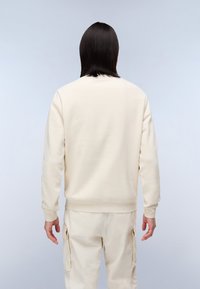 Persoon met steil zwart haar, gekleed in een gebroken witte sweatshirt en bijpassende cargobroek, staande met de rug naar de camera tegen een effen lichte achtergrond.