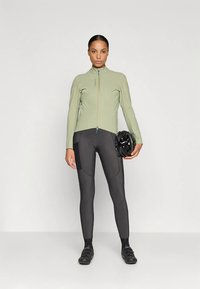 Veste de cyclisme vert clair avec un col montant et une fermeture éclair, associée à des leggings noirs ajustés. Le mannequin tient un casque noir, mettant en avant un design sportif.