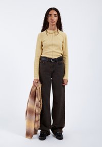 Long-sleeve yellow ribbed top, brown wide-leg jeans, black belt, and holding a brown and beige cardigan with buttons. Black shoes.