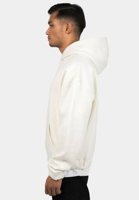 White hoodie with a kangaroo pocket, ribbed cuffs, and hem. Smooth texture, oversized design, shown from the side against a neutral background.