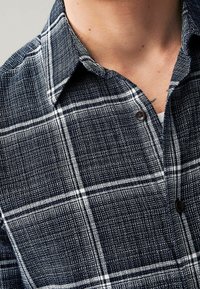 Camicia a quadri blu navy realizzata in tessuto texturizzato, con righe bianche a reticolo e bottoni scuri. Presenta un colletto classico e una zona sul petto.