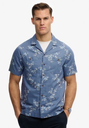 Homme portant une chemise bleue à manches courtes avec un motif floral blanc, les mains dans les poches et une montre au poignet gauche.