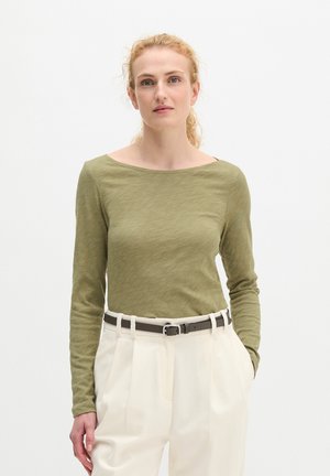 Kobieta o kręconych blond włosach, ubrana w oliwkowozielony top z długim rękawem wsunięty w białe spodnie z wysokim stanem z czarnym paskiem.