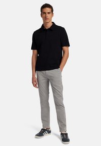 Schwarzes Poloshirt mit Kragen, schmal geschnittene graue Hose und marineblaue Sneaker mit weißen Streifen. Das Model steht lässig mit entspannten Armen.