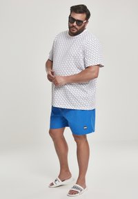 T-shirt blanche avec des lettres noires dispersées, shorts de bain bleus et sandales blanches à glissière. Le mannequin porte des lunettes de soleil noires. Textures lisses.
