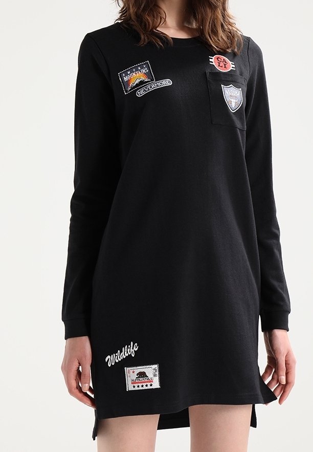 Robe noire à manches longues en tissu doux, ornée de plusieurs patchs brodés, comprenant une poche avant et des fentes latérales.