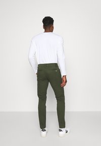 Gröna bomullsbukser med slim fit, med bakfickor och en etikett på midjebandet, i kombination med en långärmad vit skjorta och vita sneakers.