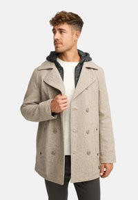 Cappotto beige doppiopetto con finitura testurizzata, grandi rever e fodera interna con cappuccio scuro. Presenta chiusura con bottoni e tasche laterali.