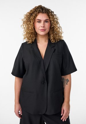 Žena s kučeravými blond vlasmi nosí čierny krátky blejzer a nohavice, ukazujúc kvetinové tetovanie na ľavom predlaktí.