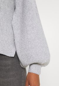 Grå stickad tröja med ribbat mönster, med volumetriska, pösiga ärmar och ett figursytt ribbat manchett. Fållen är något asymmetrisk.