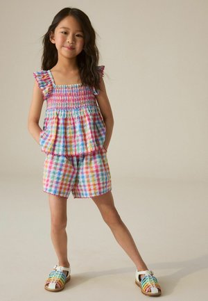 Jeune fille debout, les mains dans les poches, portant un haut à carreaux coloré et un short avec des sandales blanches arc-en-ciel sur fond neutre.