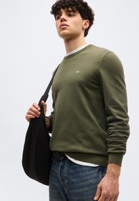 Jeune homme aux cheveux bouclés portant un pull vert olive et un jean bleu, avec un sac noir en bandoulière, regardant vers le haut.