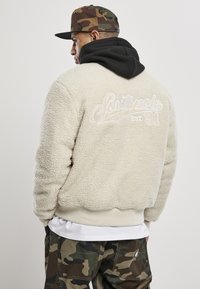 Kurtka polarowa w kolorze off-white z teksturowaną powierzchnią, z czarną bluzą z kapturem pod spodem oraz haftowanym logo na plecach.
