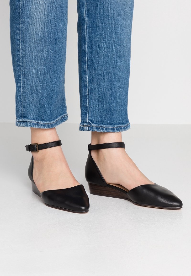 Chaussures à talons compensés en cuir noir avec un bout pointu et une bride à la cheville. Le design comprend un talon compensé bas et un dos ouvert.