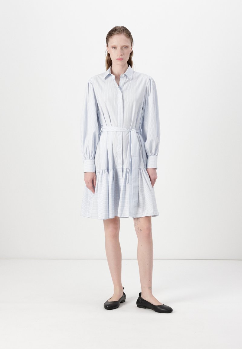 Abito camicia a righe blu chiaro con maniche lunghe, vita cinched e orlo a strati. Indossato con ballerine nere su uno sfondo semplice.