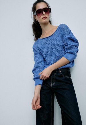 Donna con occhiali da sole grandi e scuri, un maglione blu lavorato a maglia largo e jeans scuri a vita alta, che posa davanti a uno sfondo chiaro e semplice.